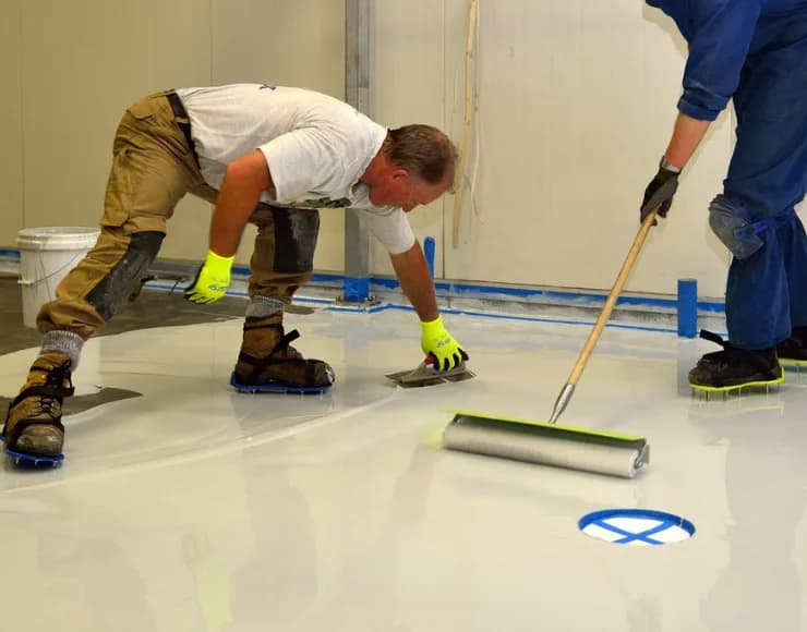 Two workers are installing or leveling a smooth, white floor surface, using a roller and a trowel, in front of a storefront related to flooring or floor leveling services.