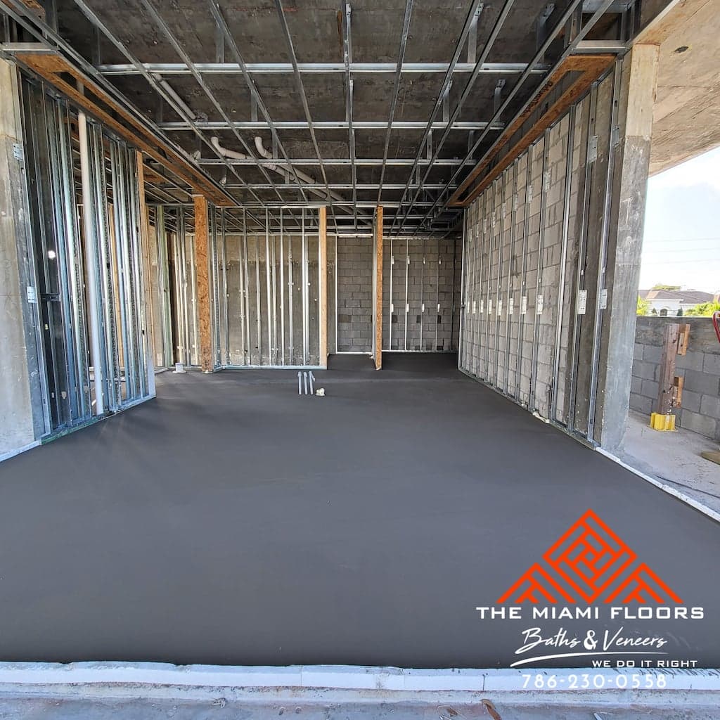 A freshly poured and smoothed concrete floor is visible inside a building under construction, with exposed metal framing and unfinished walls, indicating ongoing flooring and framing work.